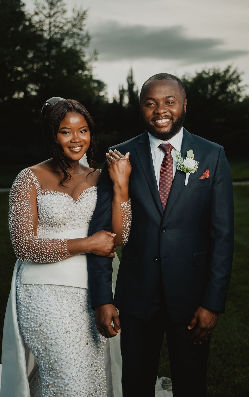 A couple poses together outdoors at dusk, wearing elegant wedding attire including a beaded gown and navy suit.