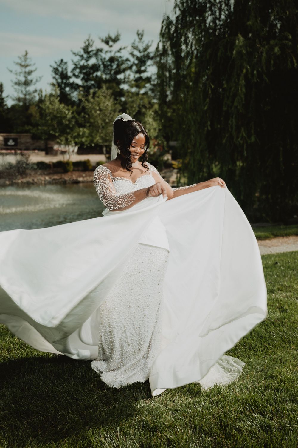 A flowing white wedding dress twirls gracefully on a lawn near a scenic waterfront.