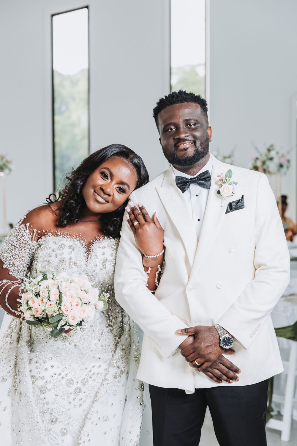 A wedding couple in elegant white attire share joyful moments in a bright indoor venue with large windows.