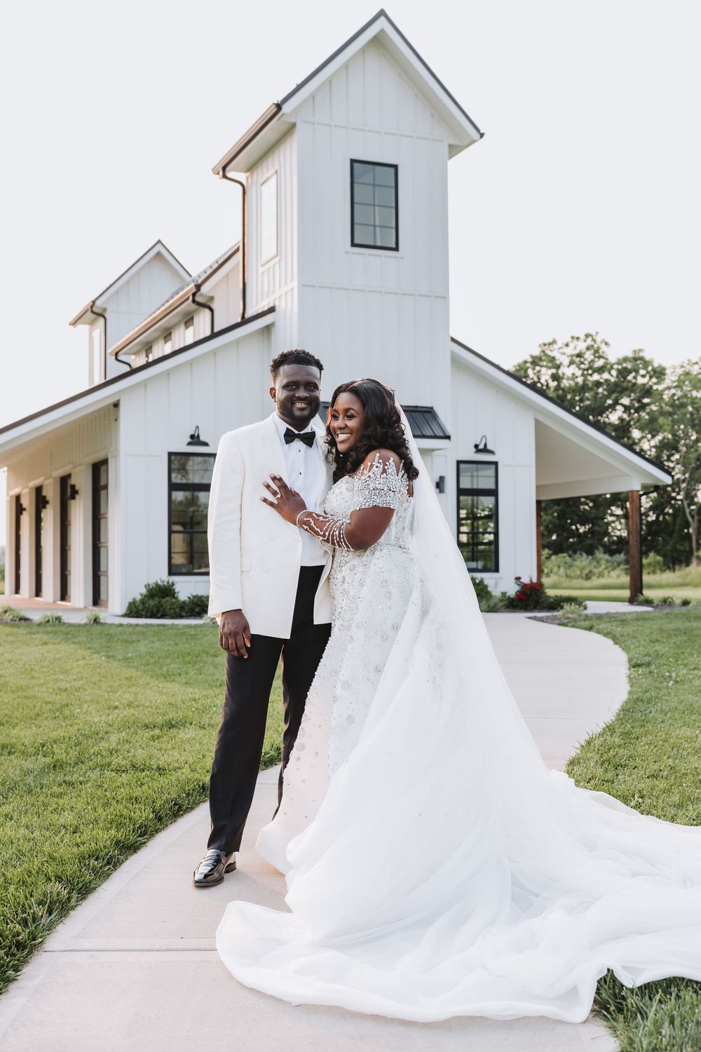 A joyful couple in wedding attire stands outside a modern white church building with a tower on a sunny day.