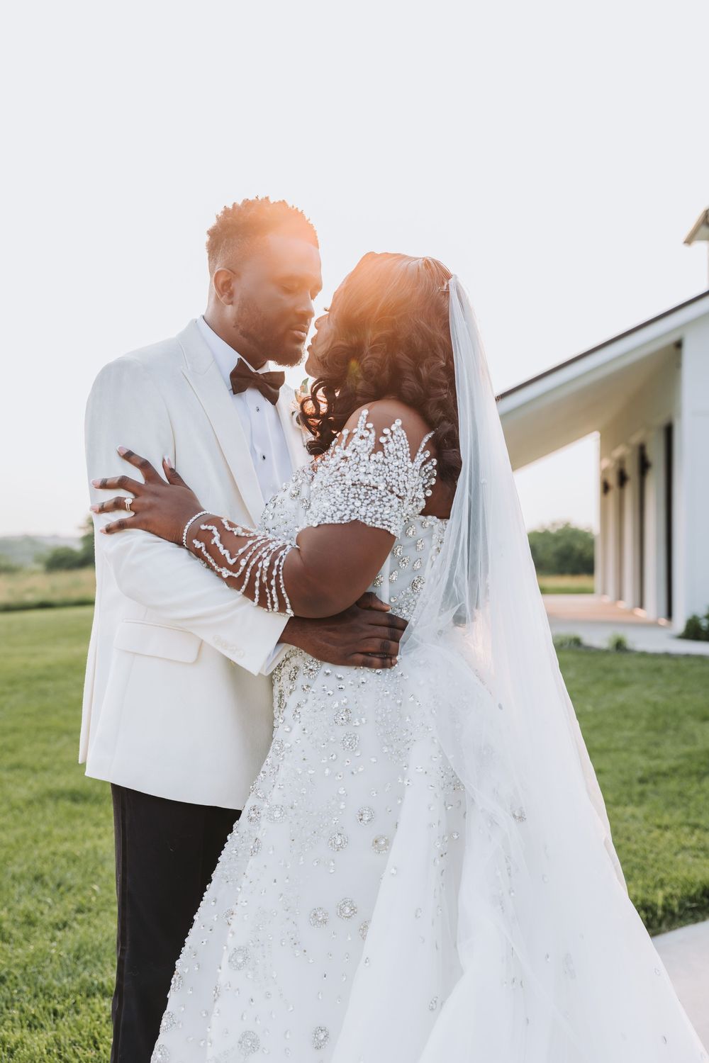 A romantic wedding couple embraces outdoors in a grassy field, with the bride wearing a white lace gown and flowing veil.
