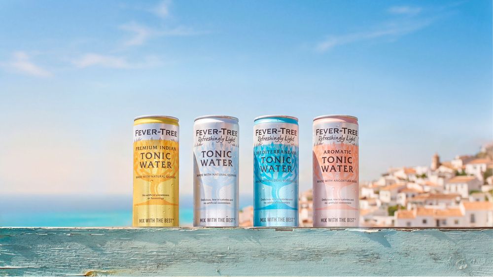 Four Fever-Tree tonic water cans displayed on a ledge with a scenic coastal town and blue sky backdrop.