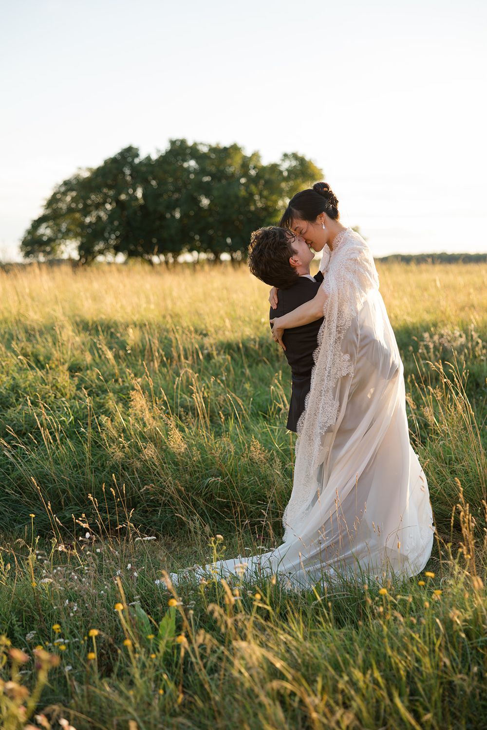 A romantic wedding moment captured in a golden field at sunset with tall grass and trees in the background. Bröllopsfotograf Emelie Petré,