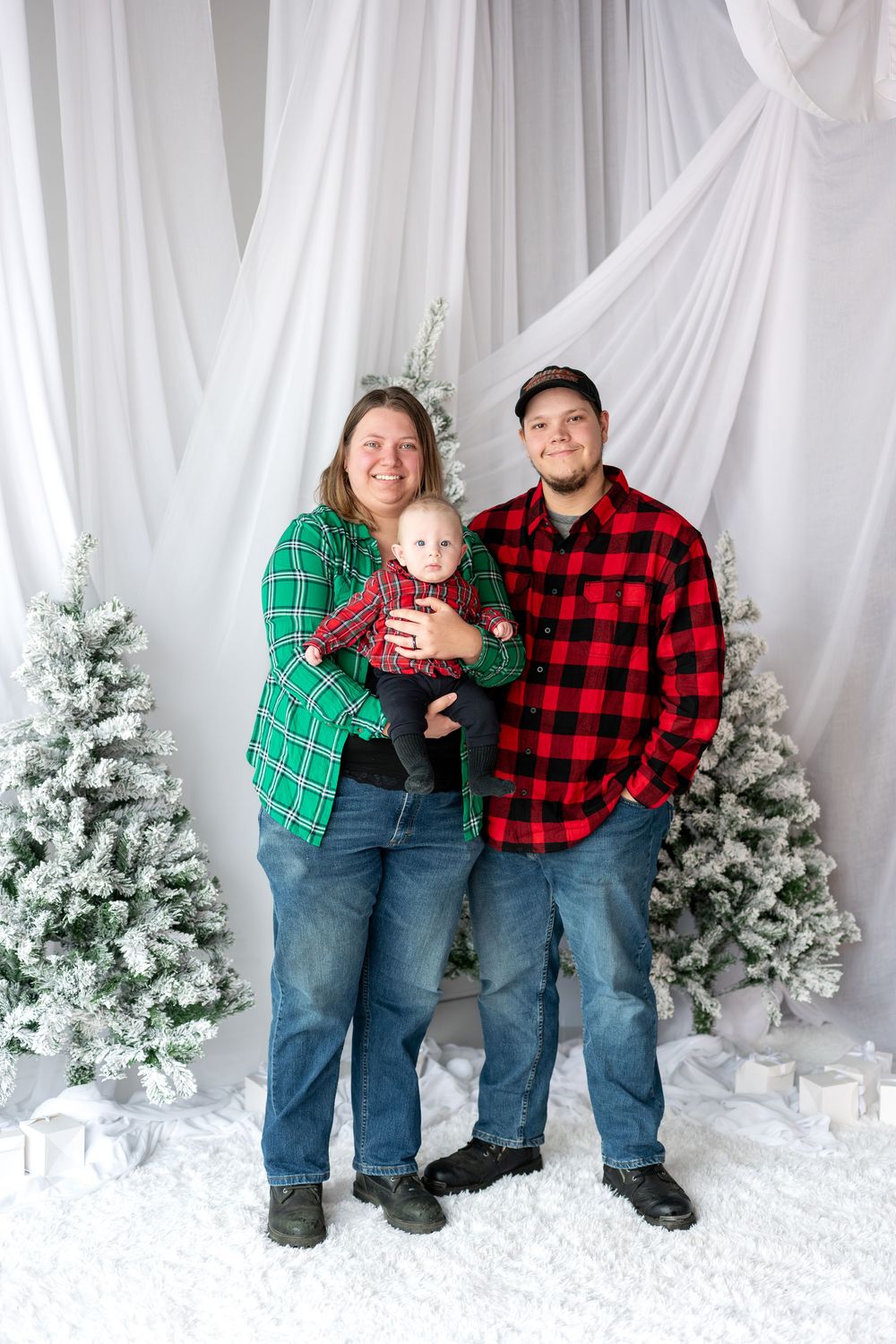 Family of three in plaid shirts and jeans posing with baby near snowy Christmas trees.