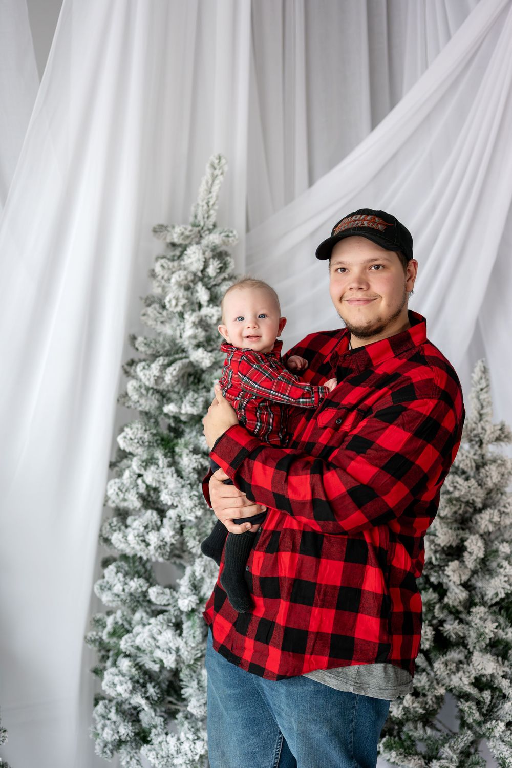 Dad in red plaid shirt and cap holding baby in matching plaid outfit beside snow-flocked Christmas trees.