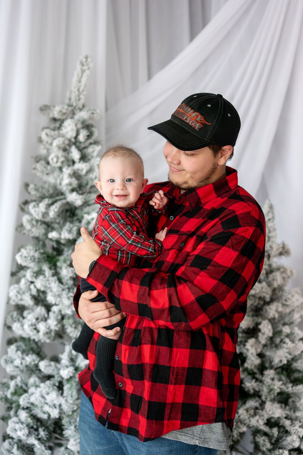 Father in red buffalo check shirt gazing lovingly at his smiling baby during a Christmas photo session.
