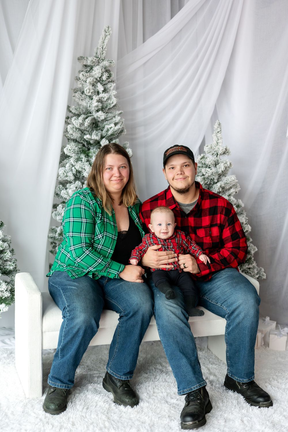 Family of three posing for Christmas photo, parents in plaid shirts with baby on white bench near snowy trees.