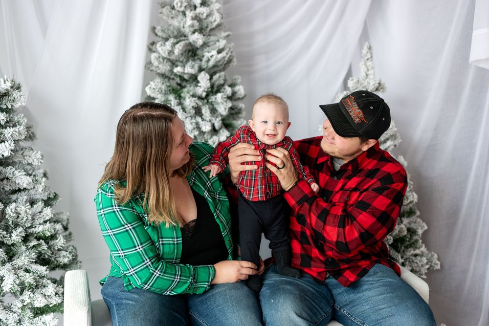 Young couple in matching plaid shirts holding baby up between snowy Christmas trees in studio.