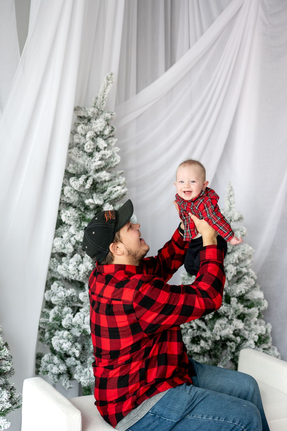 Father in red plaid shirt lifting laughing baby high in front of snow-flocked Christmas trees.