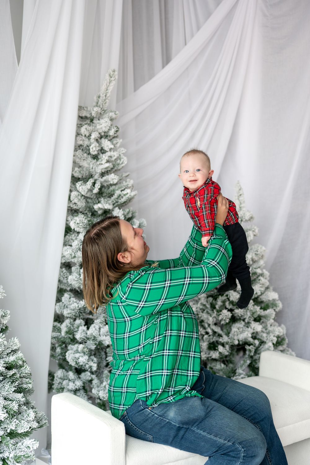 Mother in green plaid shirt lifting smiling baby above her head near decorated Christmas trees.