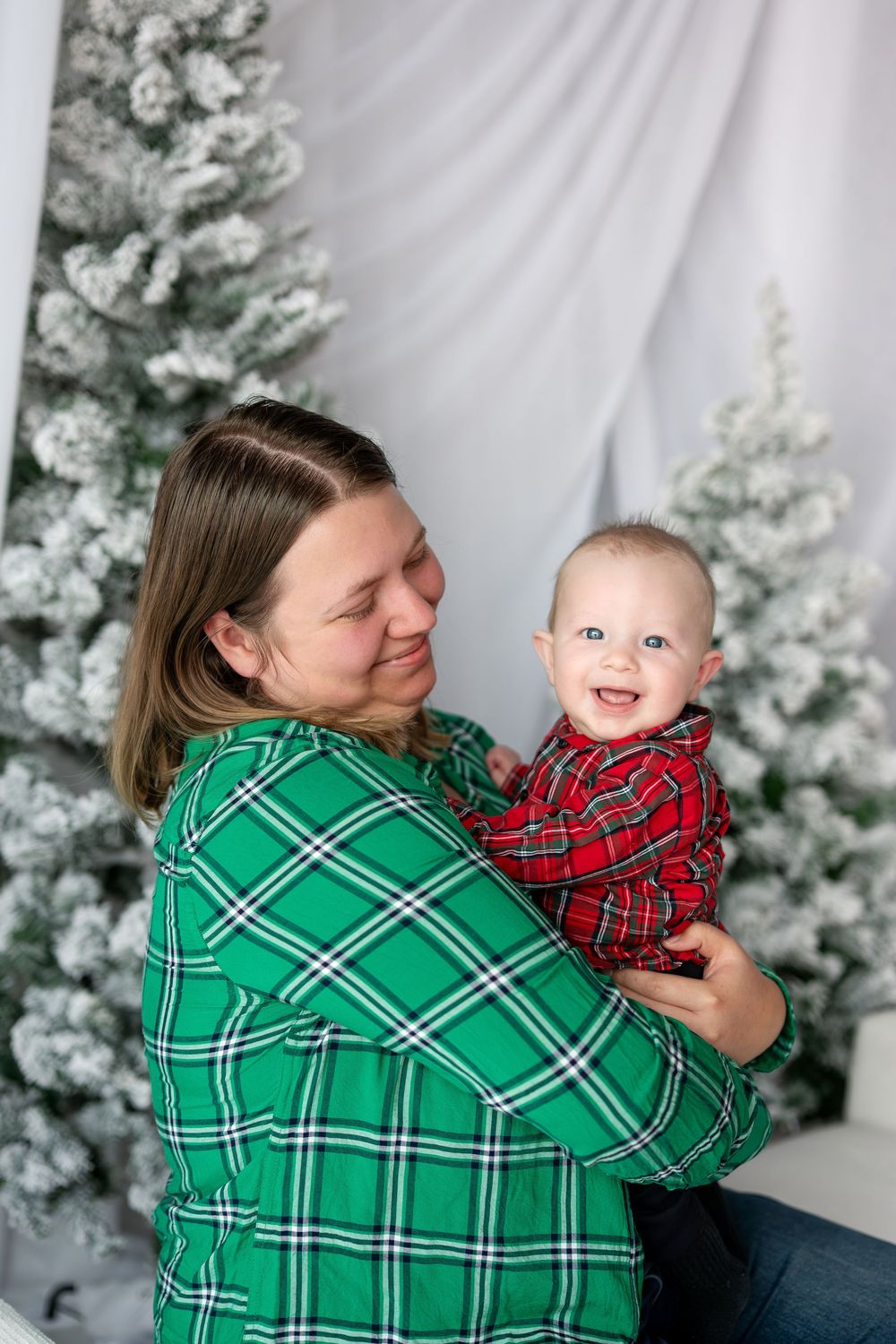 Smiling baby in red plaid outfit being held by mother in green plaid shirt near Christmas trees.
