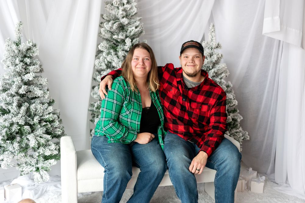 Couple in coordinating green and red plaid shirts sitting together on white bench for Christmas photo.