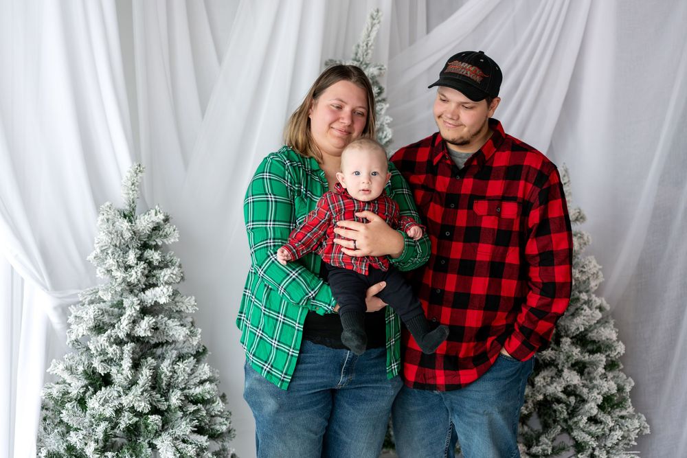 Family of three in matching plaid shirts posing together during a festive Christmas photo session with snowy trees.