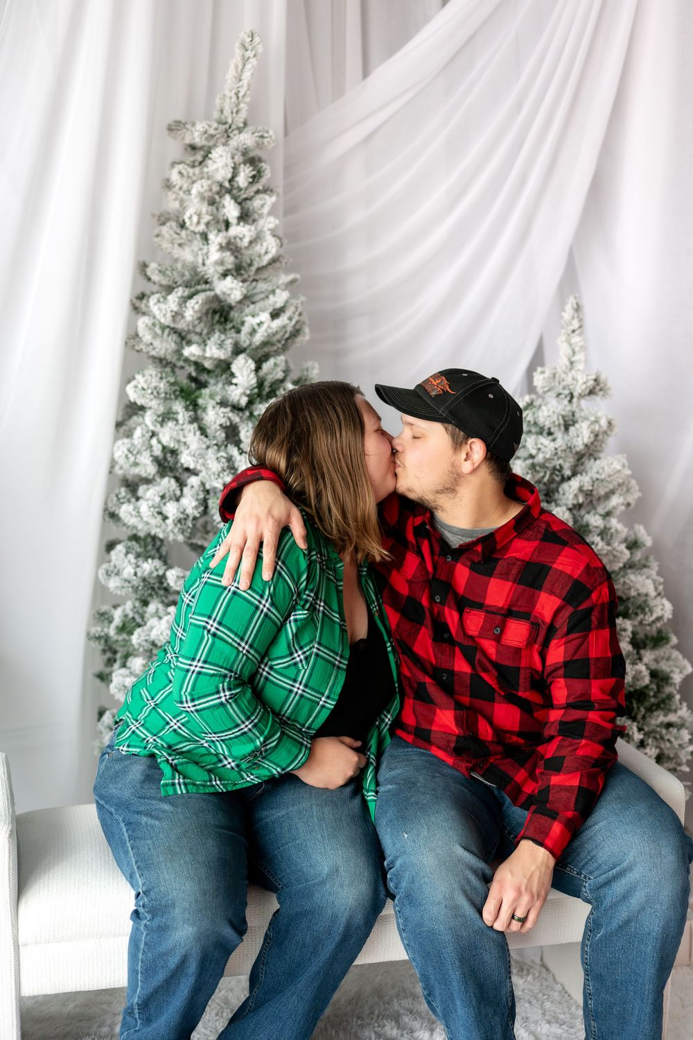 Couple sharing a kiss on white bench surrounded by snow-flocked Christmas trees in studio setting.
