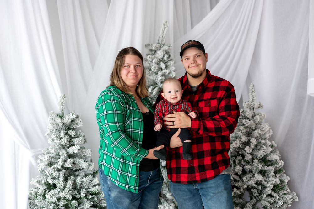 Smiling family with baby posing in front of snow-flocked Christmas trees in coordinating red and green plaid outfits.