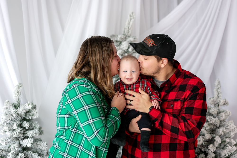 Parents in plaid shirts kissing their baby's cheeks during a Christmas family photo session with snowy trees.