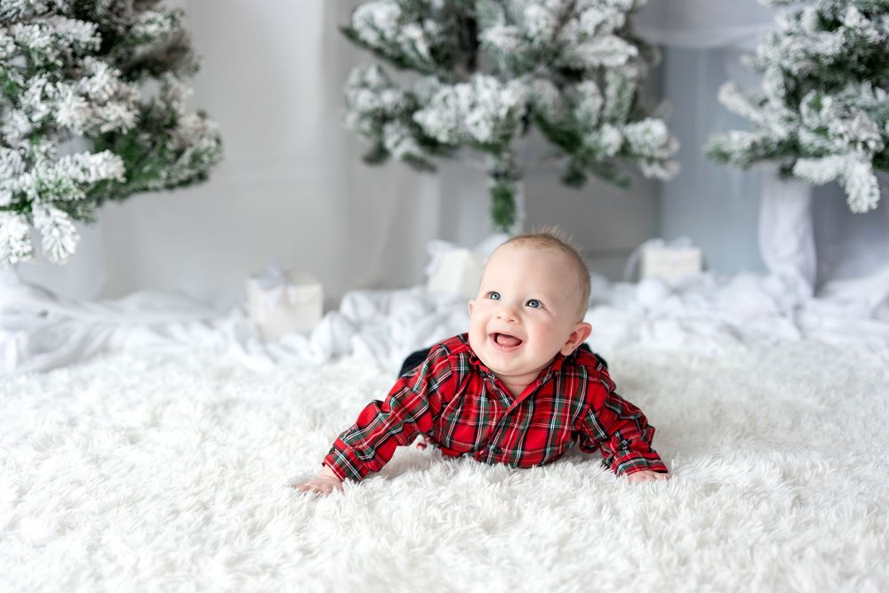 Adorable baby in red plaid shirt lying on white fluffy rug surrounded by snow-flocked Christmas trees.