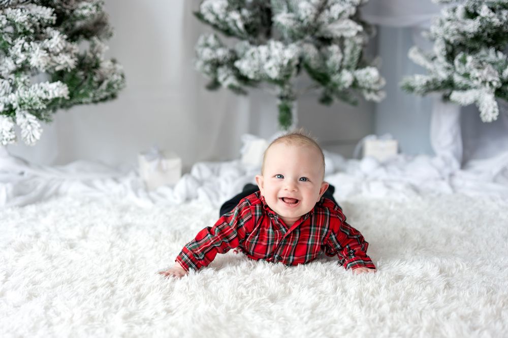 Happy baby in red plaid outfit smiling while doing tummy time on white carpet near snowy Christmas trees.