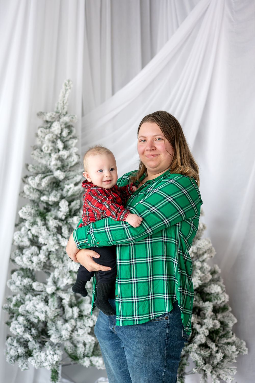 Mother in green plaid shirt holding her smiling baby during a Christmas mini session with flocked trees.