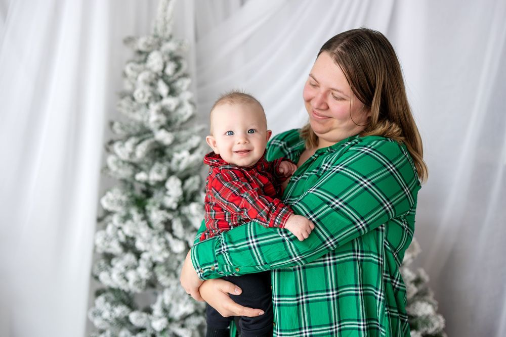 Mom in green plaid shirt cradling her baby boy near a snow-flocked Christmas tree in a studio setting.