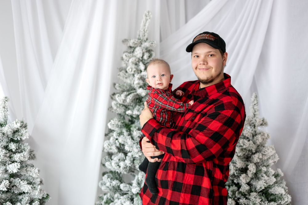 Father in red buffalo plaid shirt holding his infant during a Christmas photo session with snowy trees.