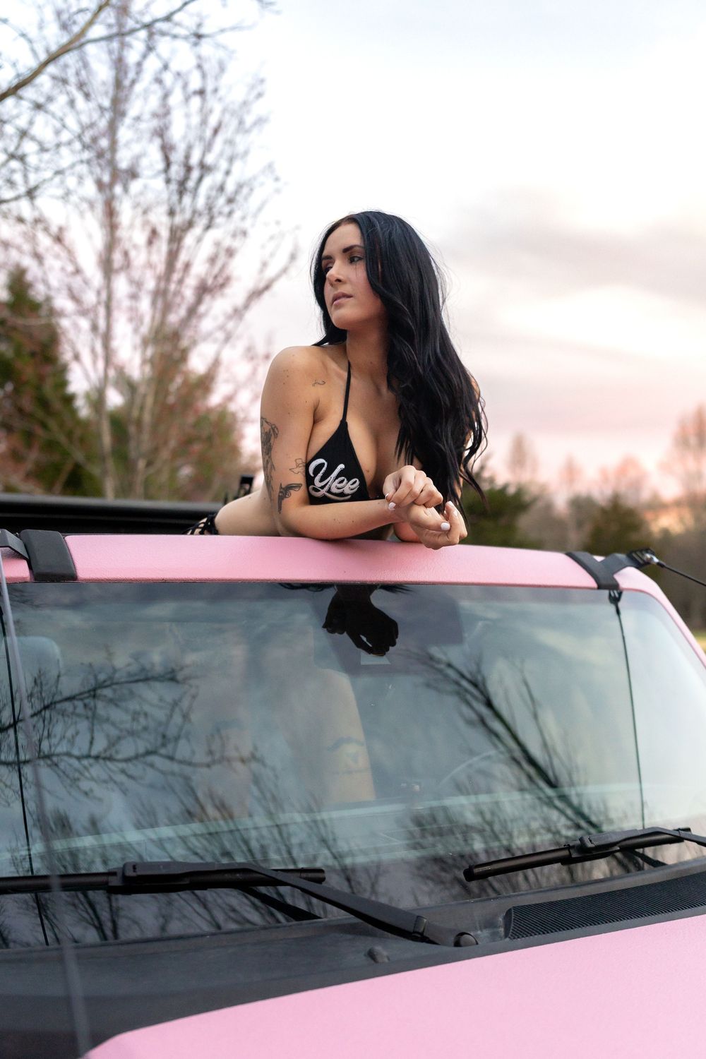 Model poses on pink car roof against sunset sky and bare trees in artistic photoshoot.