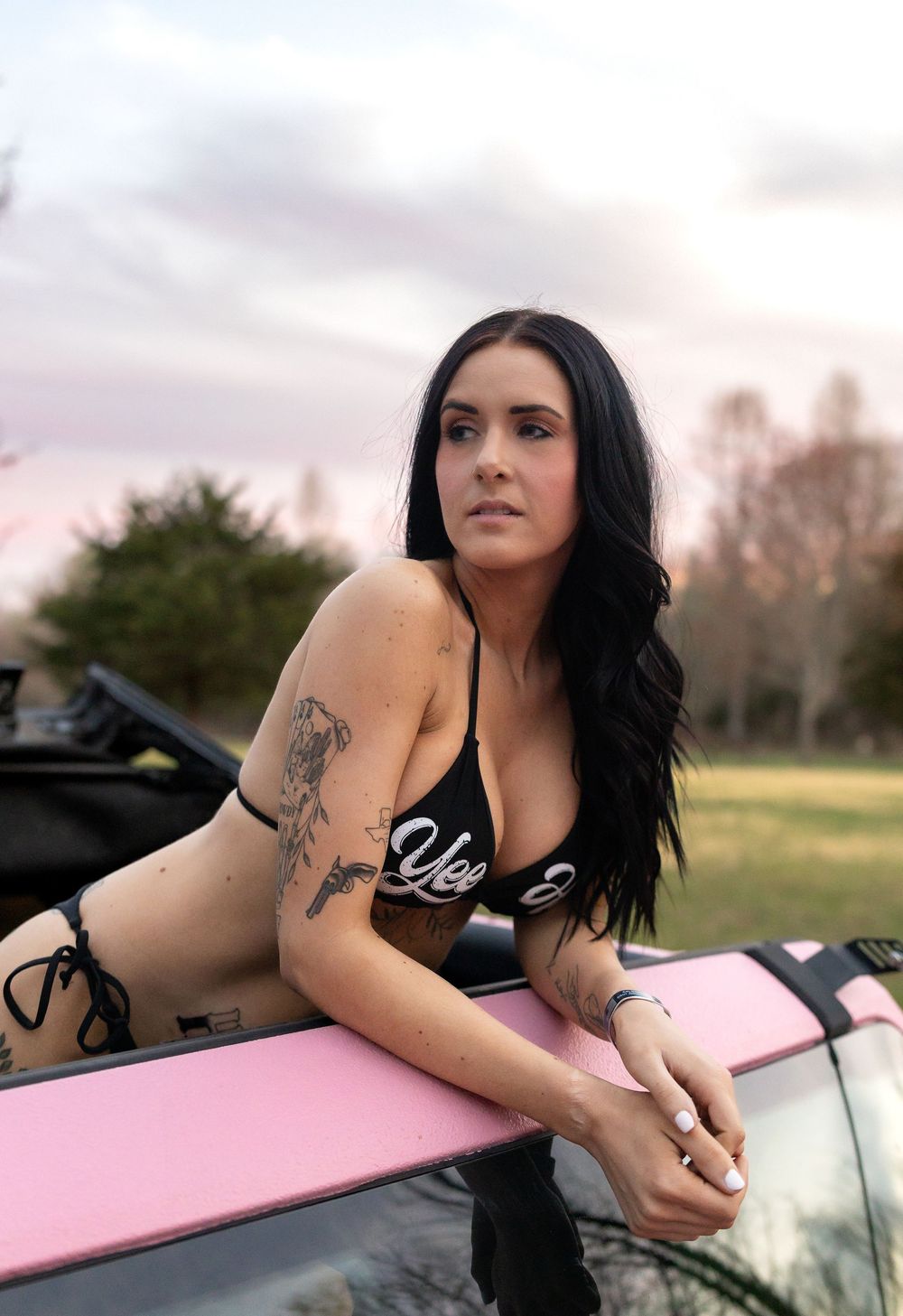 Woman with long dark hair and tattoos leans on a pink surfboard at sunset in an outdoor setting.