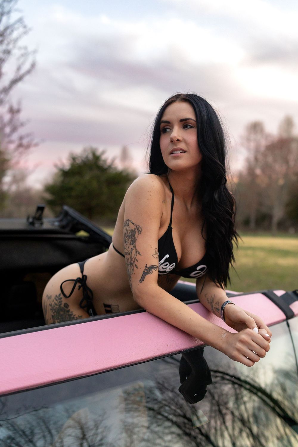 Woman with dark hair and tattoos poses confidently on vehicle during golden hour photo session.