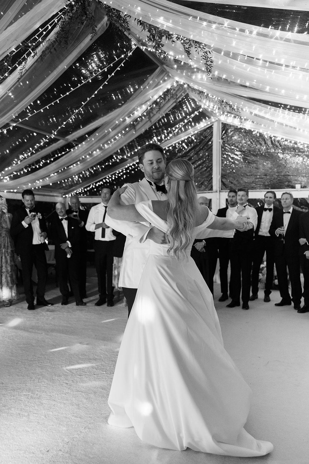 A couple shares their first dance at a wedding reception under twinkling lights and a white tent canopy.
