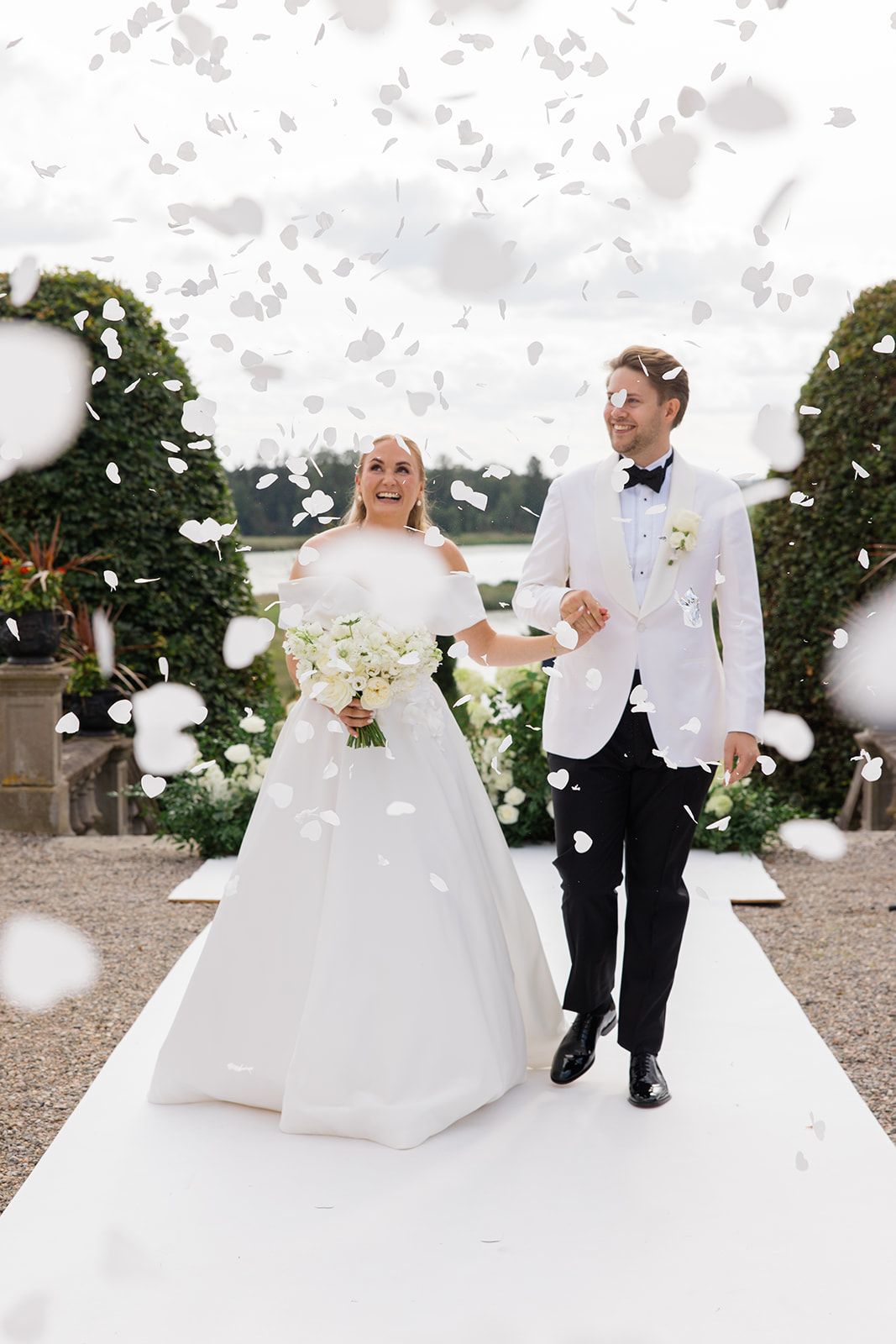 Couple in wedding attire playfully tossing white feathers in the air during outdoor photo session.