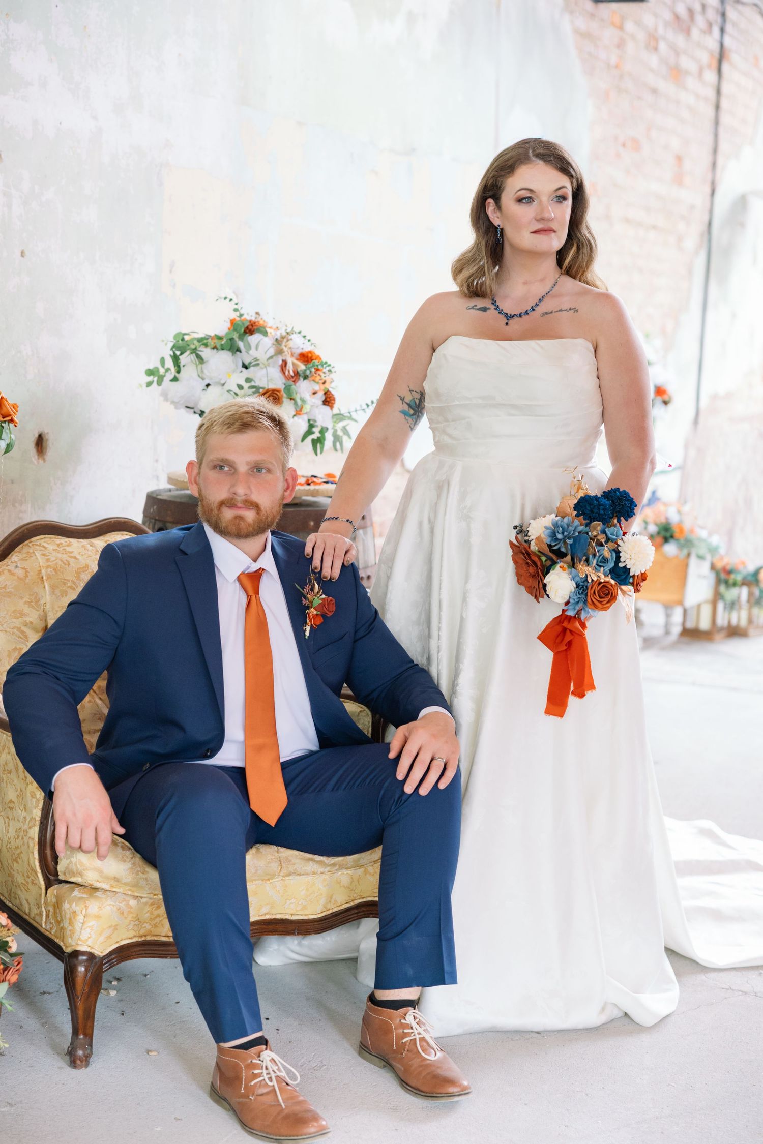 Stylish wedding couple poses on vintage sofa with navy suit and orange tie details for elegant autumn portraits.