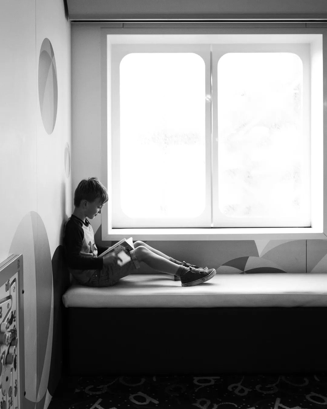Boy sitting at a desk reading by a bright window in a black and white room.