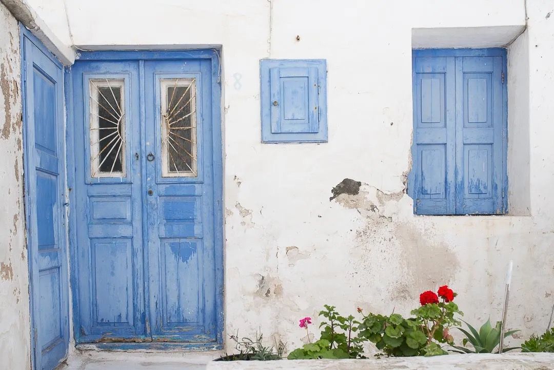 A whitewashed Greek home with bright blue doors and windows, decorated with potted flowers.