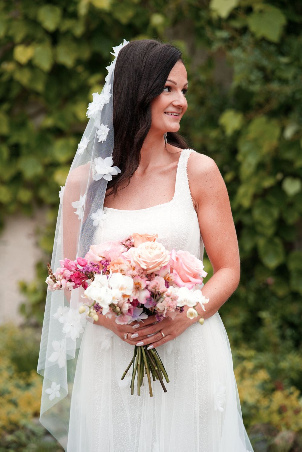 Bride in white dress holding pink and peach floral bouquet against green garden backdrop.