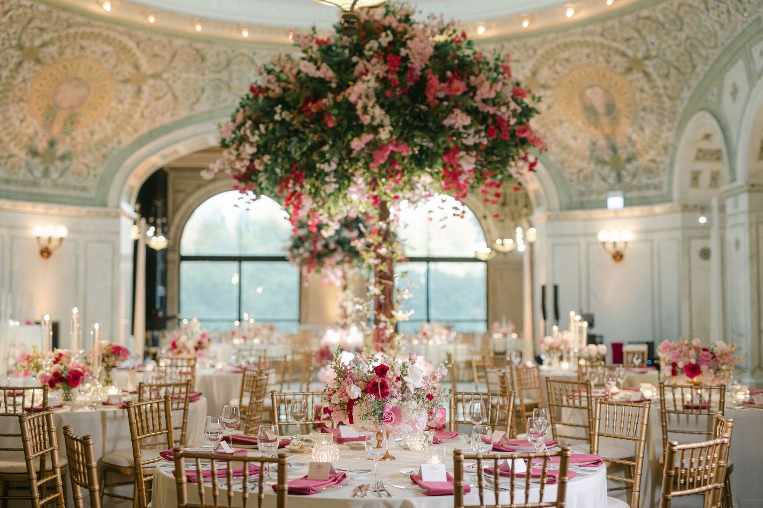 Elegant wedding reception hall decorated with pink floral arrangements and gold chairs in a grand ballroom setting.
