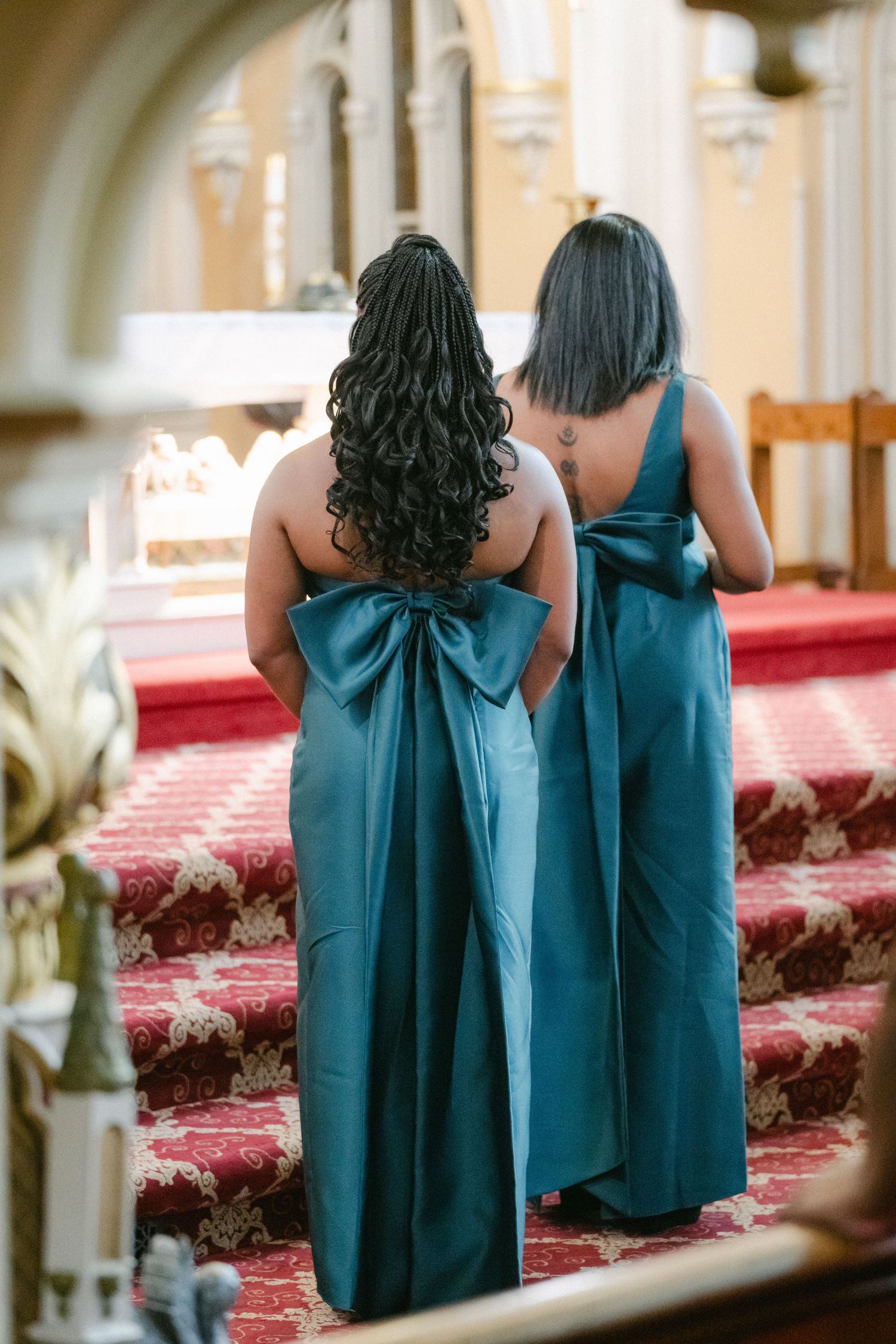 Three bridesmaids in teal dresses with large bows stand in a church aisle with red carpeting.