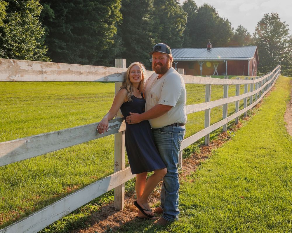 A couple embraces on a sunny day by a white wooden fence stretching across a green rural field.