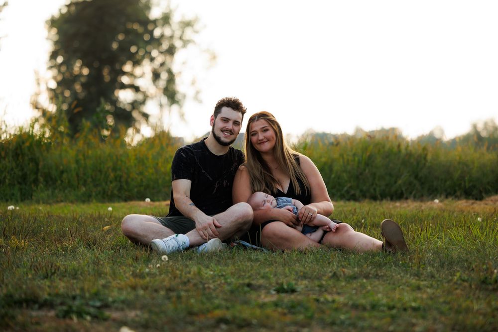 A couple and their baby relaxing together on a grassy hill during a golden sunset photo session.