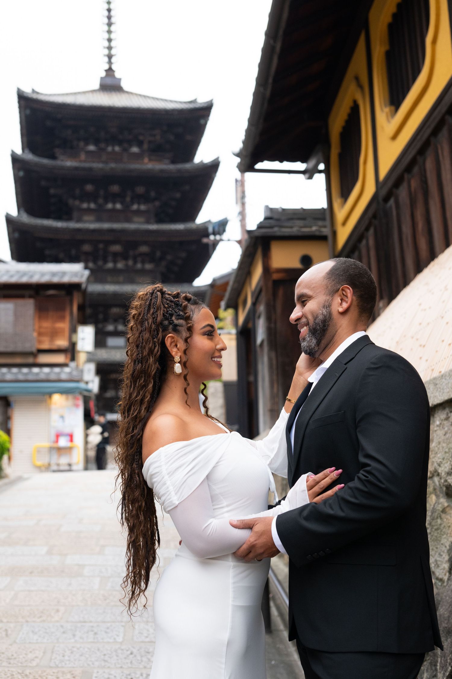 Couple smiles at each other near Yasaka Pagoda, woman in white off-shoulder gown, Kyoto Japan.