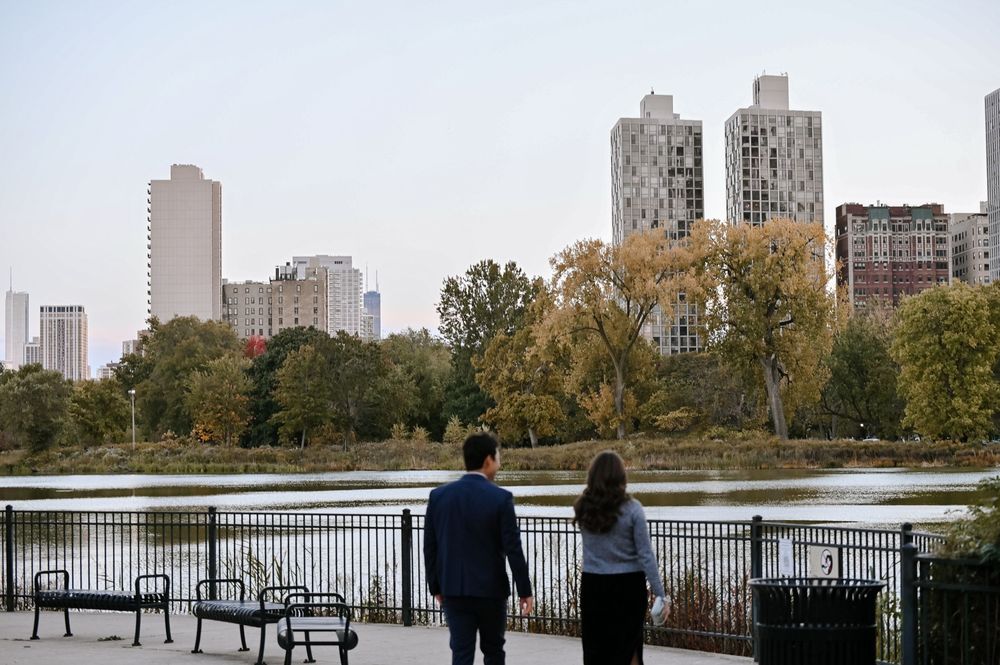 People walking along a lakefront path with autumn trees and city skyline in the background.