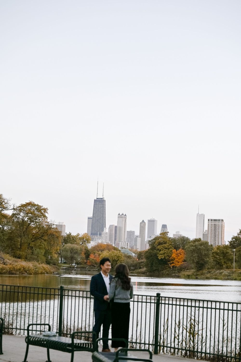 A couple sits on a bench overlooking a lake with Chicago's skyline visible in the autumn afternoon.