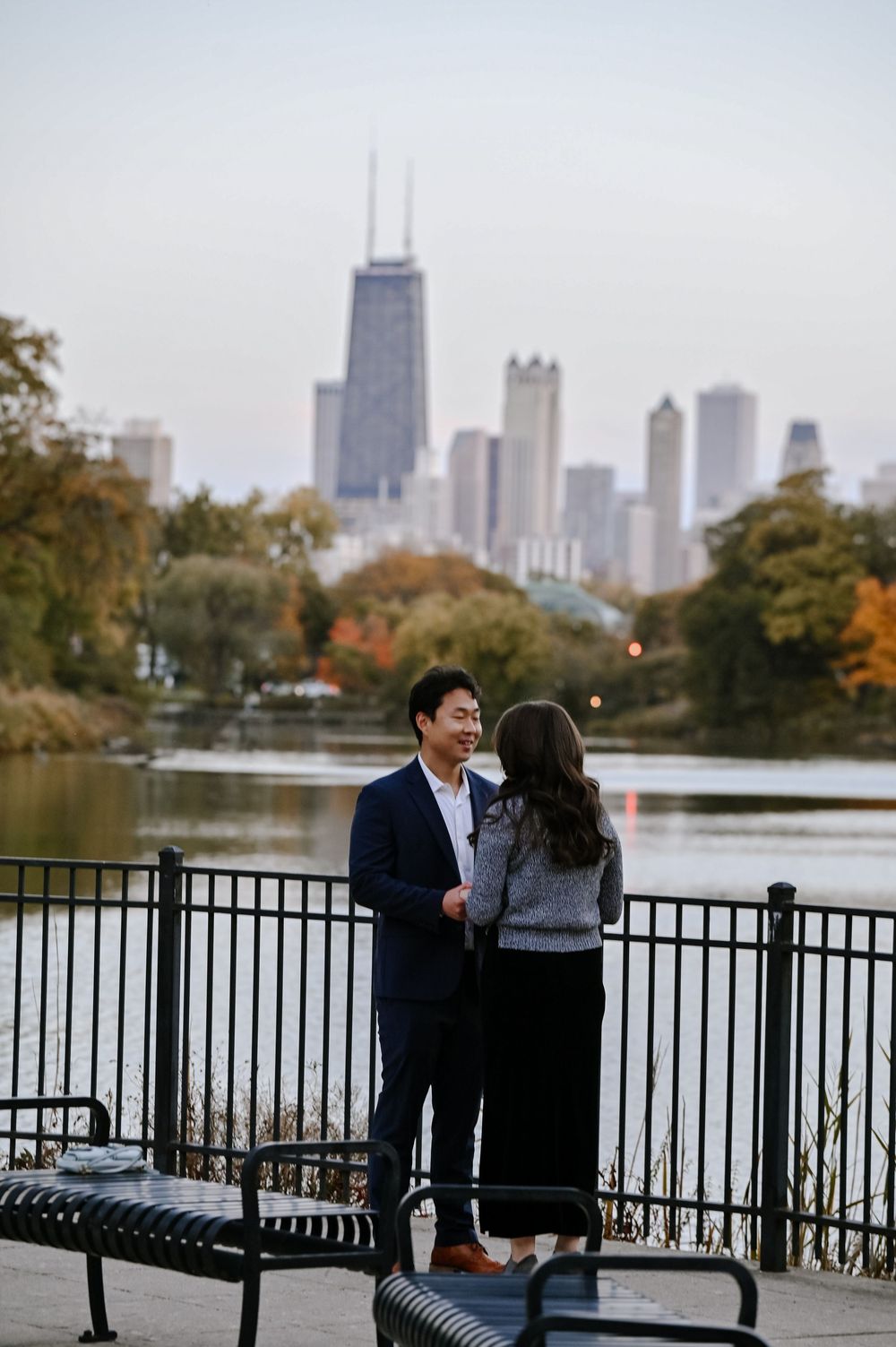 A couple shares a romantic moment by a lake with the Chicago skyline and fall foliage in the background.