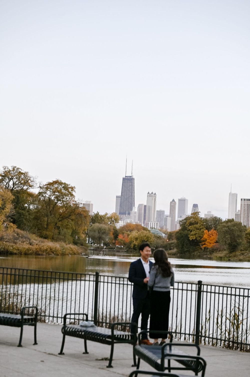 A couple sits on park benches overlooking a scenic lake with Chicago's skyline visible in the autumn landscape.