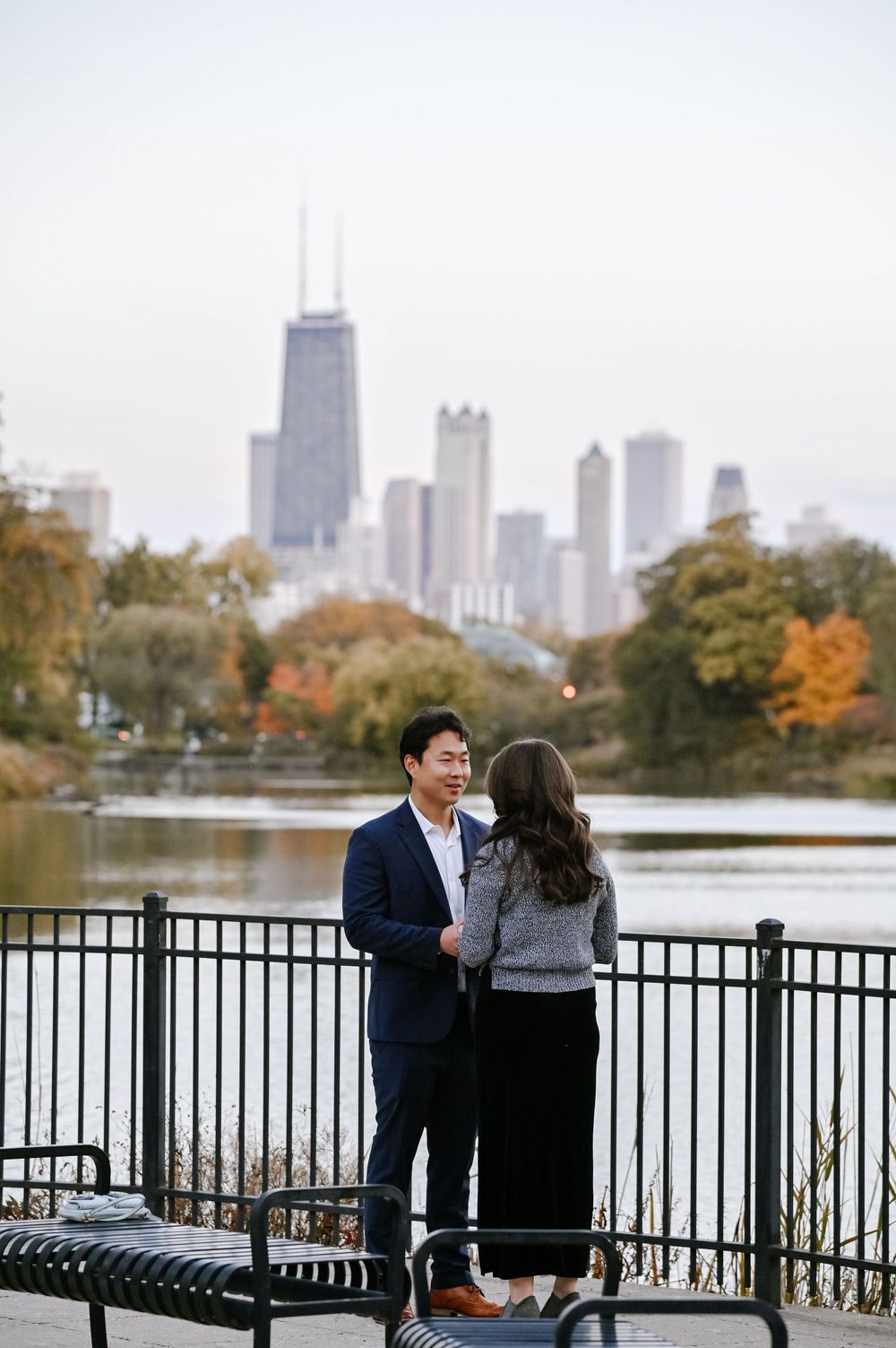 A couple shares a romantic moment by a lakefront railing with the Chicago skyline visible in the autumn afternoon.
