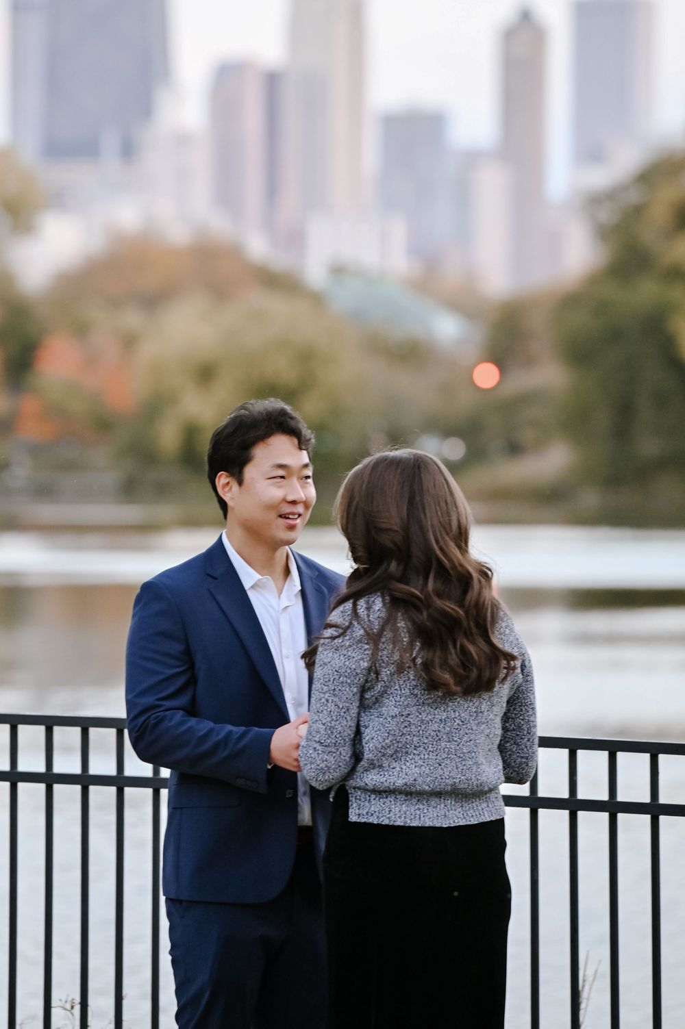 A couple shares an intimate moment along a waterfront railing with autumn trees and city skyline in the background.