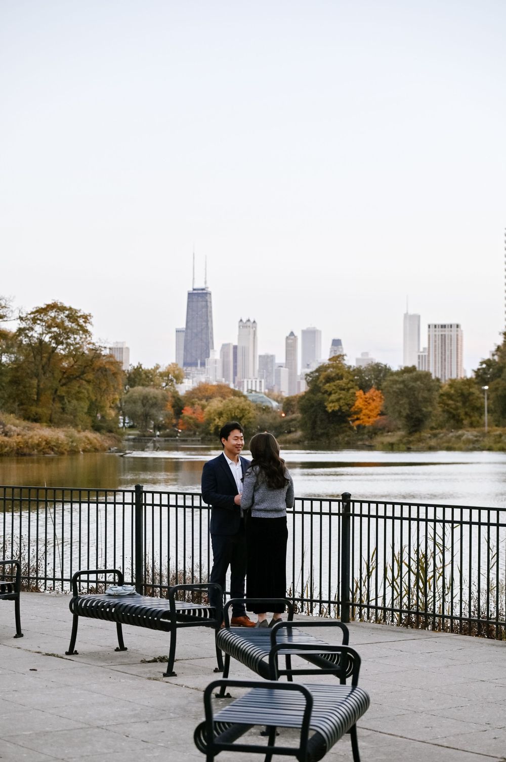 Two people share a moment on park benches by a lake with the Chicago skyline visible in fall.