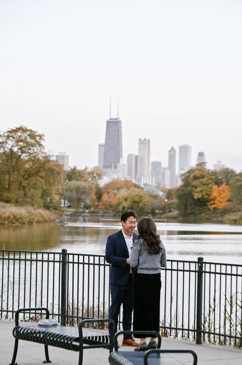 Two people stand on a bench overlooking a lake with Chicago's iconic skyline and autumn trees in view.