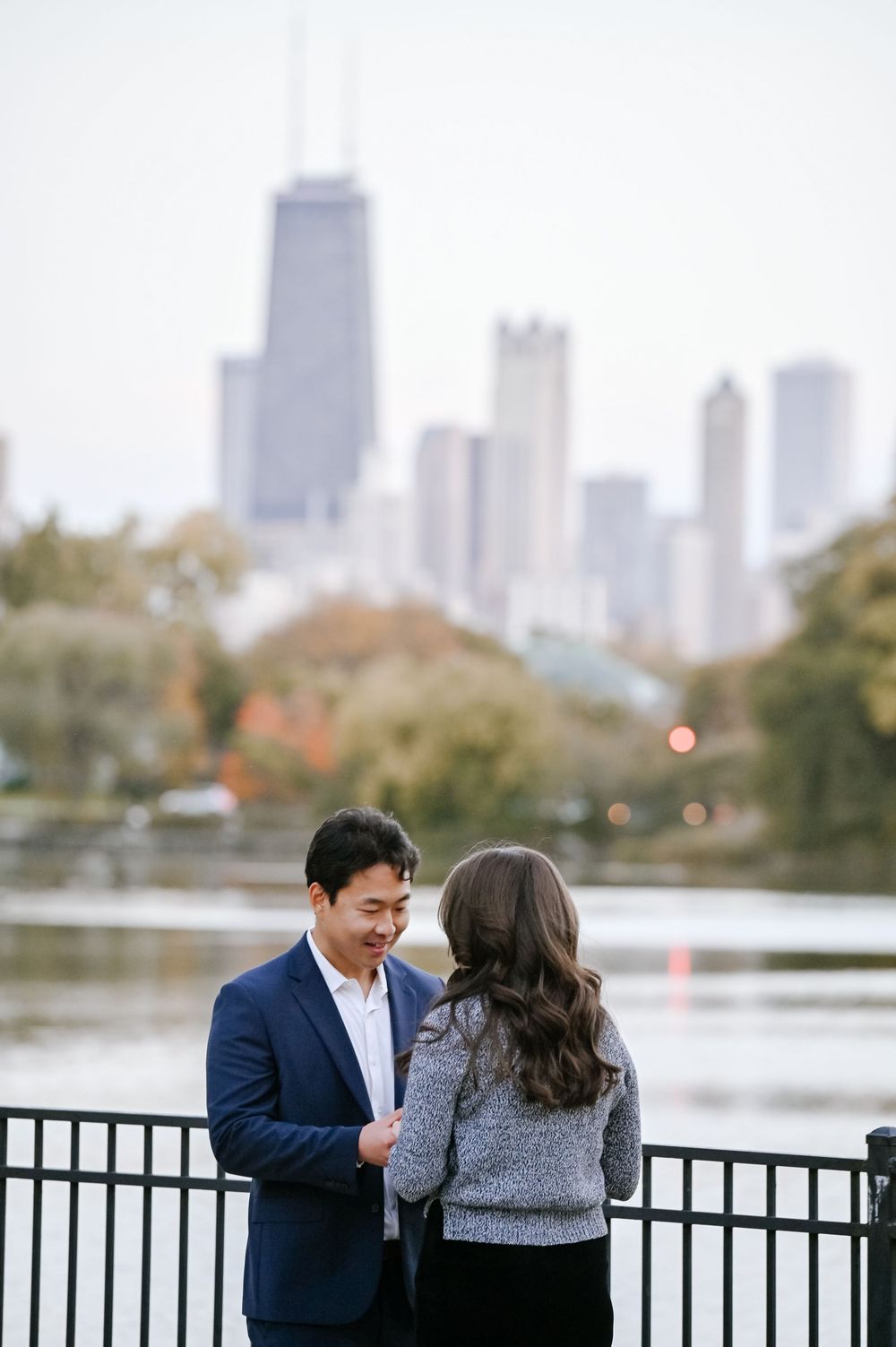 A couple shares a romantic moment by a lakeside railing with a city skyline in the background during autumn.