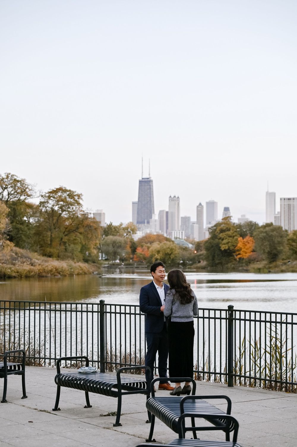 Park benches line a waterfront path with fall foliage and downtown Chicago skyscrapers in the background.