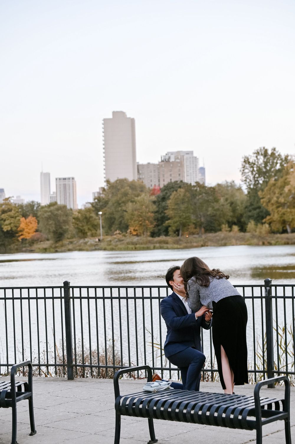 A scenic lakeside view shows benches and fall foliage with Chicago's urban skyline in the distance.
